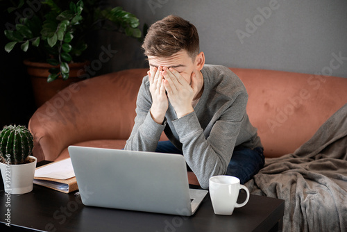 Feeling tired. Frustrated young  man looking exhausted and covering his face with hands while sitting at laptop his home working place