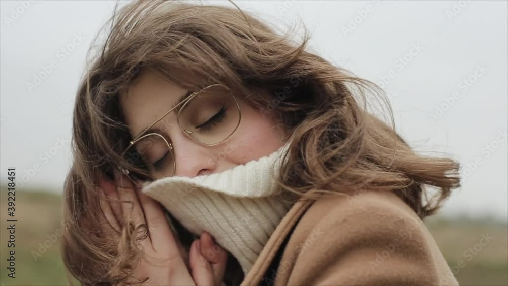 Portrait of a sensual young woman standing against the sky and pulling the collar of a sweater over her nose with closed eyes Portrait of a sensual young woman standing against the sky and pulling the collar of a sweater over her nose with closed eyes