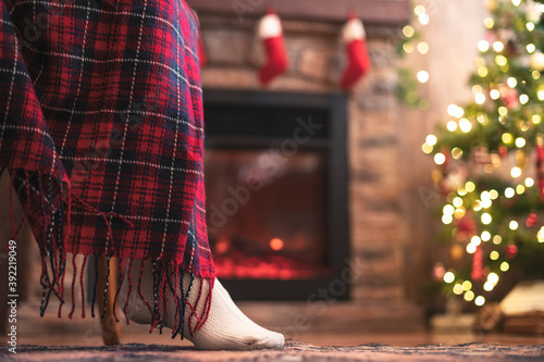 Bottom view of disabled woman legs in knitted winter socks sitting on armchair covered plaid near christmas tree and fireplace. Bottom view