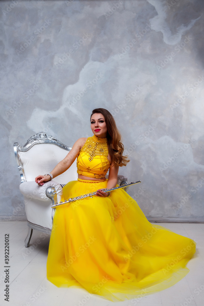 Flute player with flute instrument. Pretty actress in yellow dress with beauty Golden hair on chair. Cute young woman with flute. Classic female musician on texture wall background. Copy space