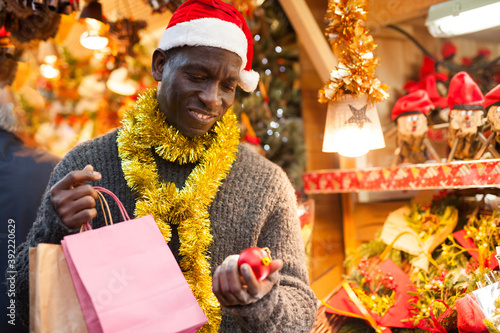 African-American guy wearing warm sweater and Santa hat walking through street Christmas fair in festive mood, choosing handmade decorations