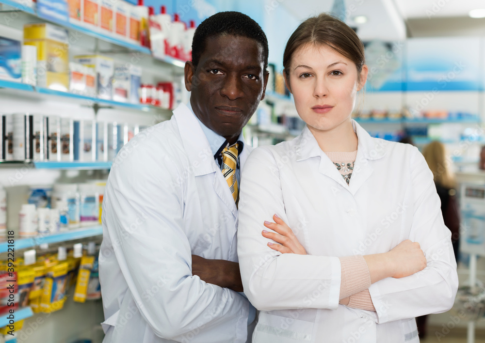Fototapeta premium Two cheerful confident pharmacists standing on background with shelves of medicines in pharmacy