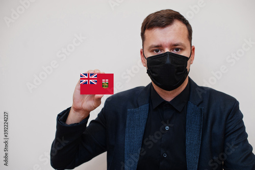 Man wear black formal and protect face mask, hold Manitoba flag card isolated on white background. Canada provinces coronavirus Covid country concept. photo
