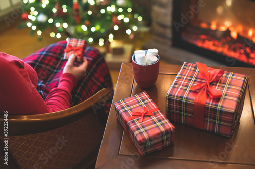 Alone woman covered plaid with cup of cocoa with marshmallows sitting and relaxation on armchair near fareplace and christmas tree after finishing pakking gift boxes for fami photo