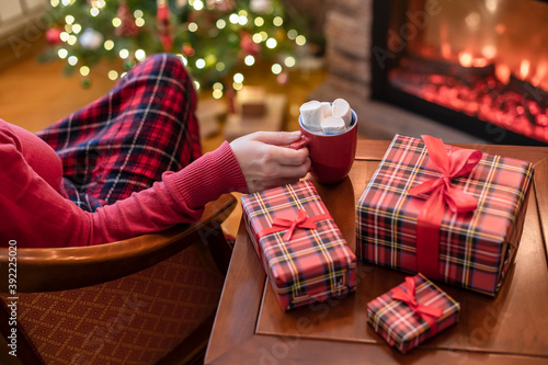 Alone woman covered plaid with cup of cocoa with marshmallows sitting and relaxation on armchair near fareplace and christmas tree after finishing pakking gift boxes for fami