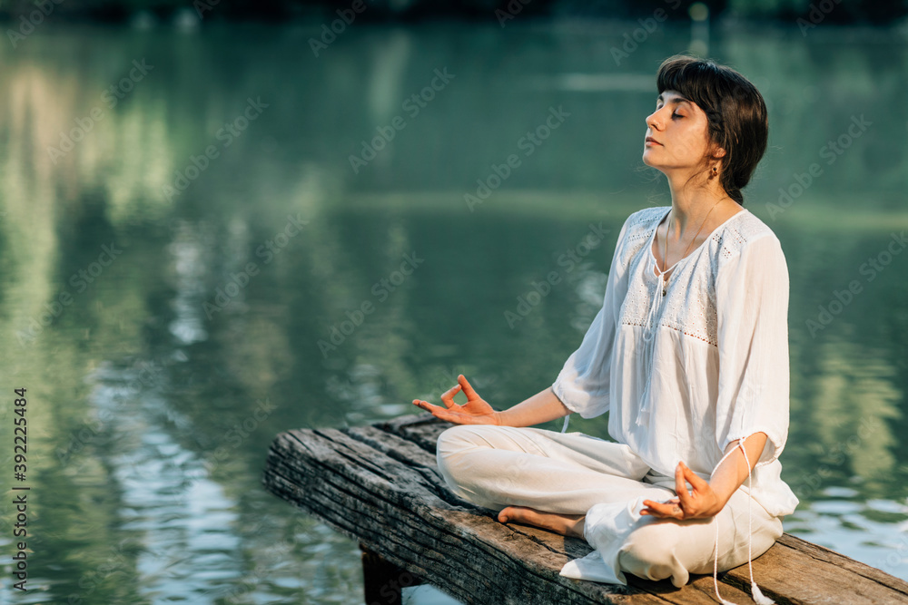 Yoga Retreat. Peaceful Young Woman Sitting in Lotus Position and Meditating by the Lake Yoga Retreat. Peaceful Young Woman Sitting in Lotus Position and Meditating by the Lake