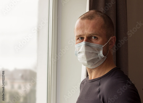 A man in a mask looks out the window, The concept of quarantine and loneliness.