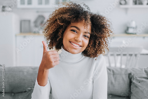 Happy young woman with trendy kinky hairstyle and dark skin smiles showing thumbs-up against modern white kitchen close view photo