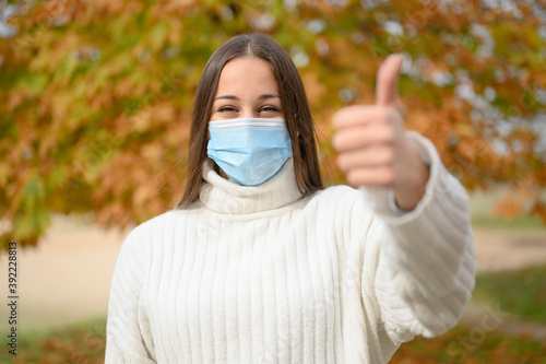 Front view portrait of a happy woman with protective mask gesturing thumbs up in the street