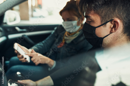 Man and woman sitting in a car using smartphones wearing the face masks to avoid virus infection and to prevent the spread of disease in time of coronavirus
