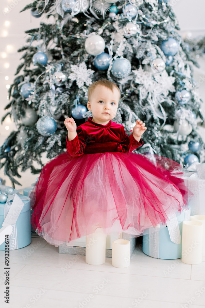 Christmas and New Year celebration. Cute little girl in a beautiful red dress sitting near the Christmas tree with silver and blue decorations.