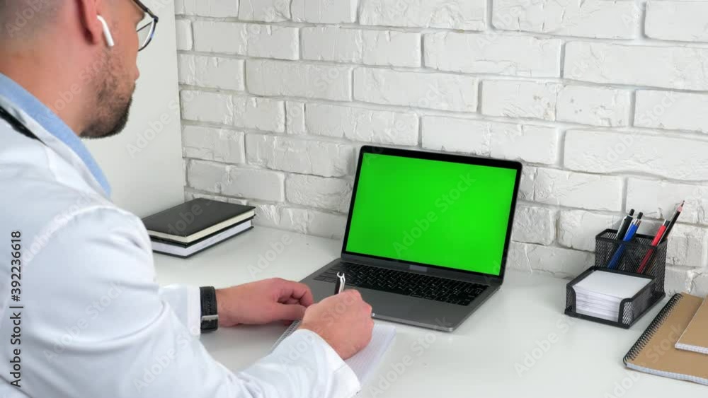Man doctor wears medical coat wireless earphones sits on chair at desk in hospital office looking green screen laptop computer listens patient client writes symptoms in notebook, social distancing Man doctor wears medical coat wireless earphones sits on chair at desk in hospital office looking green screen laptop computer listens patient client writes symptoms in notebook, social distancing
