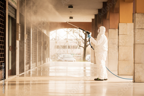 Worker in protective suit spraying disinfectant in empty arcade – public sanitation and infection control concept