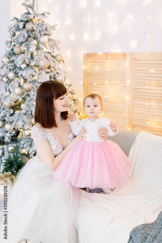Young attractive family, pretty red haired mother with cute little baby girl, sitting on the sofa, on the background of decorated Christmas tree and lights Young attractive family, pretty red haired mother with cute little baby girl, sitting on the sofa, on the background of decorated Christmas tree and lights