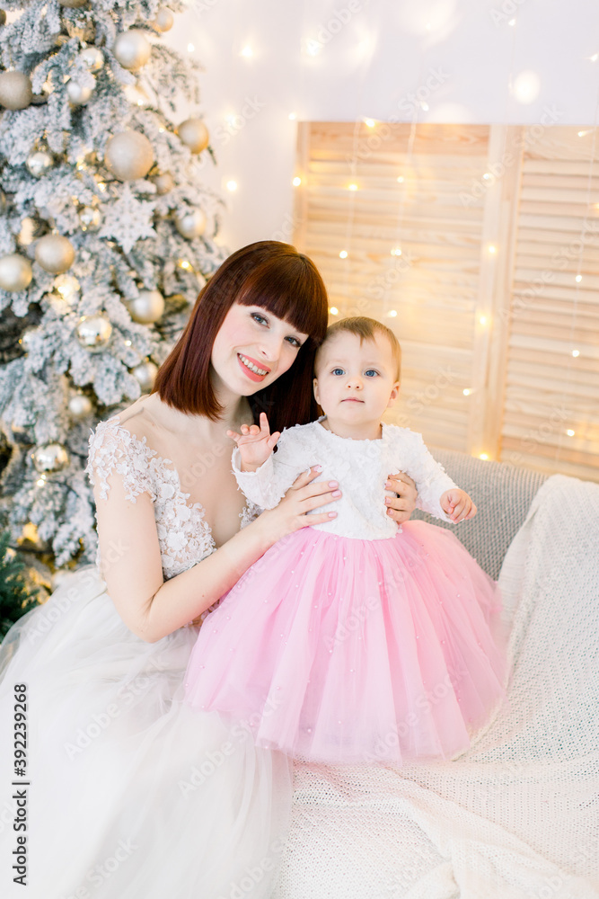 Young attractive family, pretty red haired mother with cute little baby girl, sitting on the sofa, on the background of decorated Christmas tree and lights Young attractive family, pretty red haired mother with cute little baby girl, sitting on the sofa, on the background of decorated Christmas tree and lights
