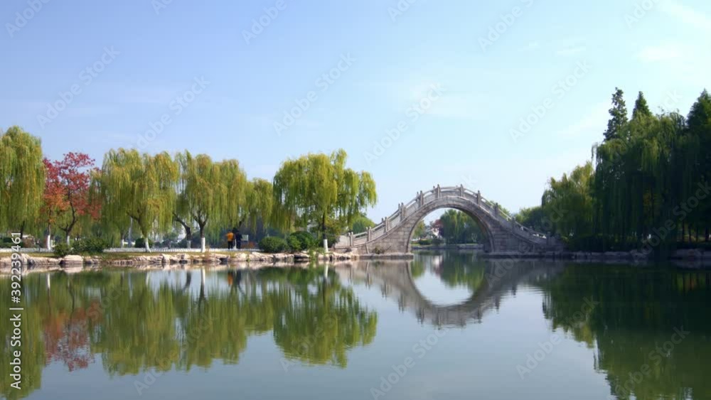 Stone arch bridge and willows in park reflected on lake, Chinese garden scene Stone arch bridge and willows in park reflected on lake, Chinese garden scene