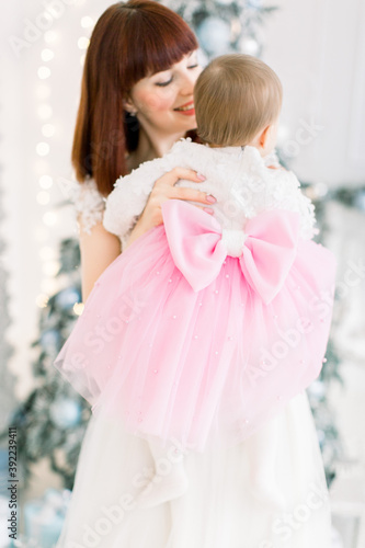 Little baby girl wearing beautiful pink and white dress with big bow on the back  enjoying time with her lovely smiling mommy  at light decorated room with Christmas tree