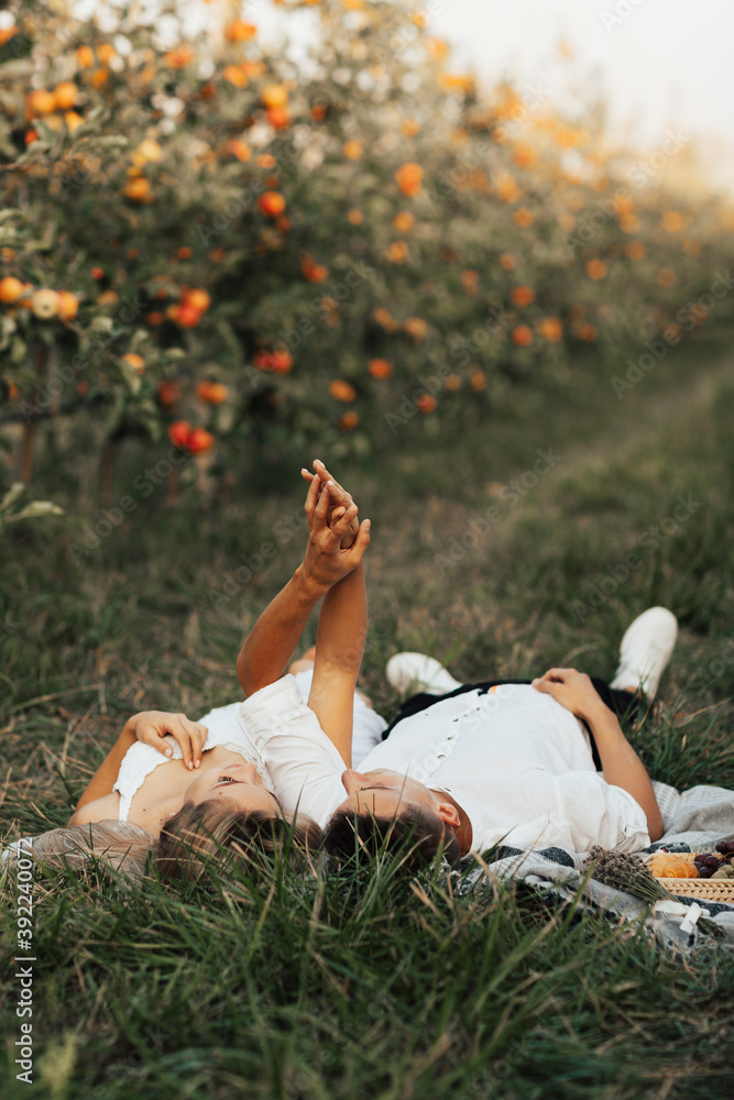 Happy couple in love is lying on the green grass in the park and enjoying the summer day together. They holding hands up and looking each other.
