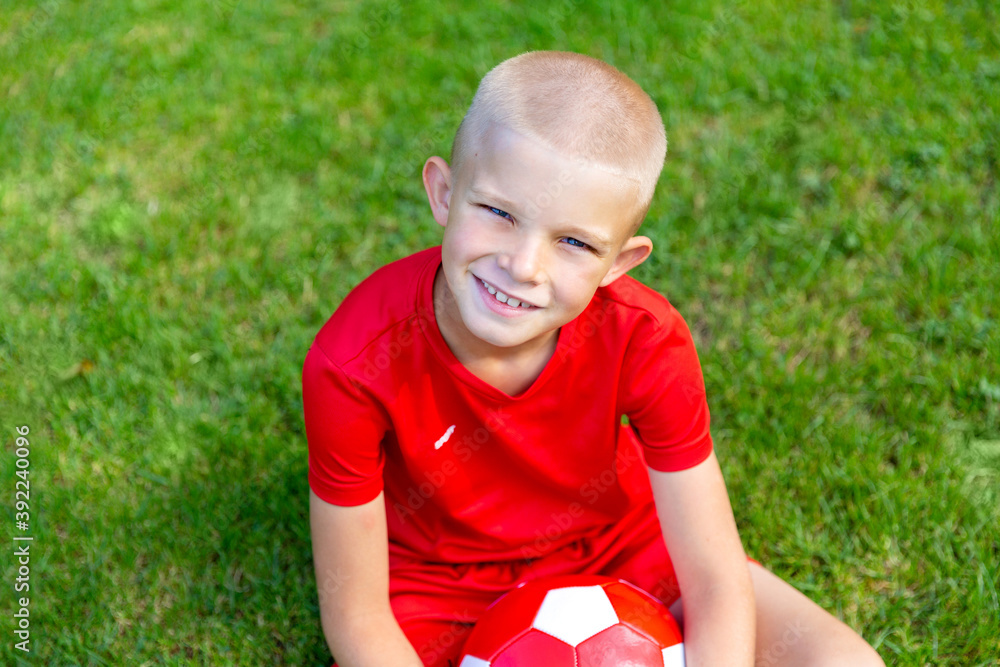 A blond, short-haired boy in a red football uniform sits on the football field and smiles at the camera A blond, short-haired boy in a red football uniform sits on the football field and smiles at the camera