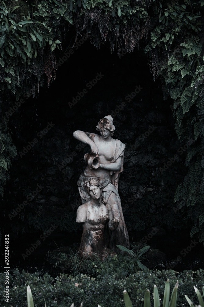 Estatua femenina en una fuente de agua rodeada de vegetación con fondo oscuro Estatua femenina en una fuente de agua rodeada de vegetación con fondo oscuro