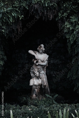 Estatua femenina en una fuente de agua rodeada de vegetación con fondo oscuro