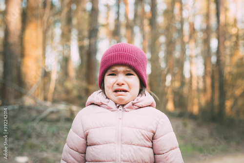 Photography a 3-year-old girl in a pink jacket and a pink hat stands in the forest and cries