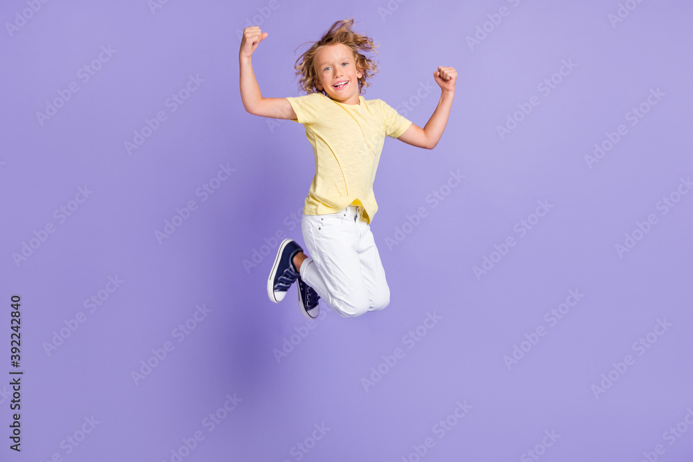 Full length photo of positive boy jump show biceps wear white yellow t-shirt pants isolated over purple color background Full length photo of positive boy jump show biceps wear white yellow t-shirt pants isolated over purple color background