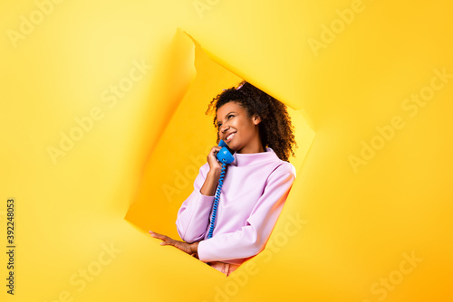 happy african american woman talking on retro telephone near hole in ripped paper on yellow background © LIGHTFIELD STUDIOS