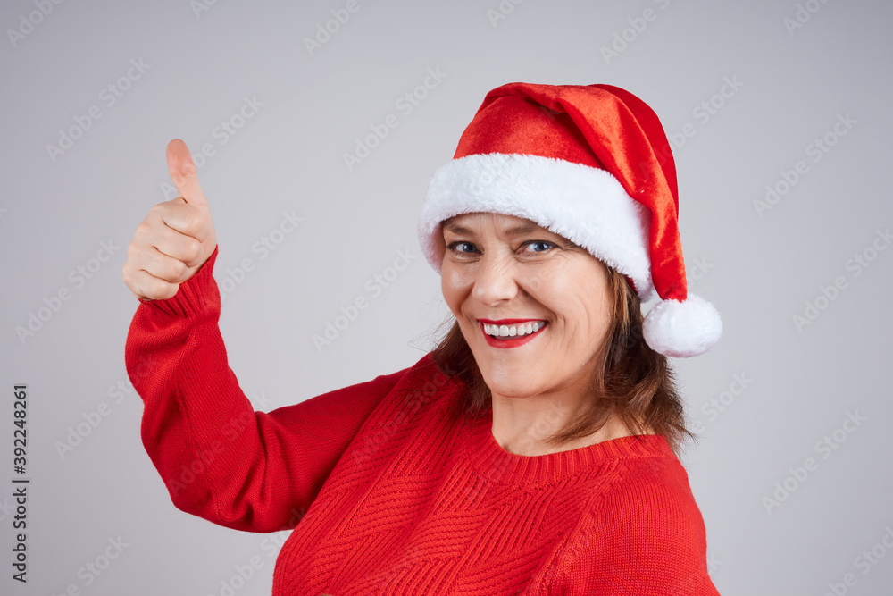 Cheerful, positive mature woman in santa hat, red sweater, holds her finger up. Concept for Merry Christmas, Happy New Year Cheerful, positive mature woman in santa hat, red sweater, holds her finger up. Concept for Merry Christmas, Happy New Year