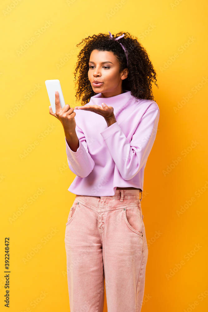 african american woman holding smartphone and sending air kiss on yellow african american woman holding smartphone and sending air kiss on yellow