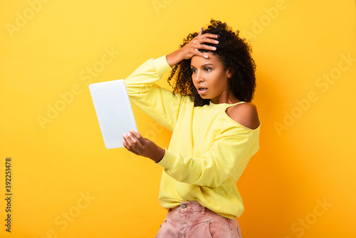 shocked african american woman looking at digital tablet on yellow