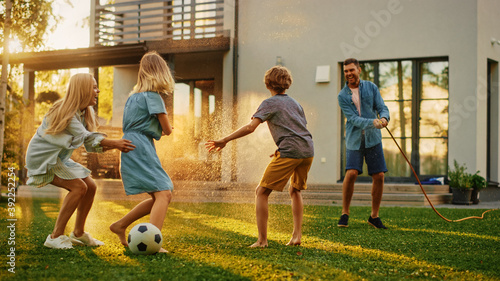 Happy Family of Four Playing with Garden Water Hose, Spraying Each Other. Mother, Father, Daughter and Son Have Fun Playing Games in the Backyard Lawn of Idyllic Suburban House on Sunny Summer Day
