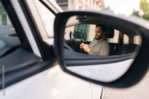 Car's side-view mirror reflecting Caucasian bearded young man sitting in car and typing online message on cell phone. Male sitting in modern vehicle and works on smartphone.