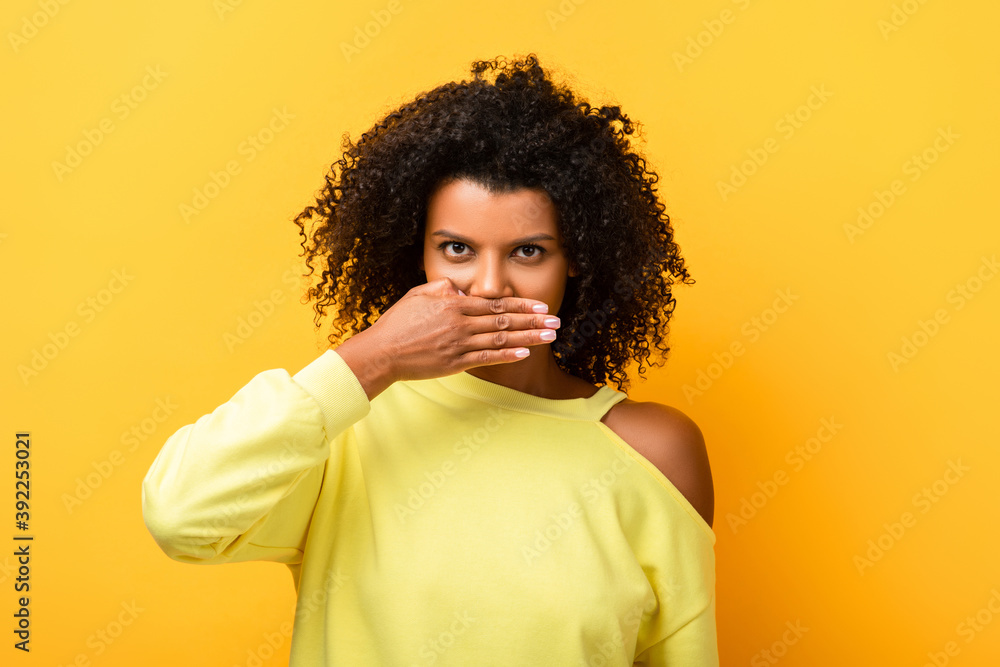 african american woman covering mouth and looking at camera on yellow african american woman covering mouth and looking at camera on yellow