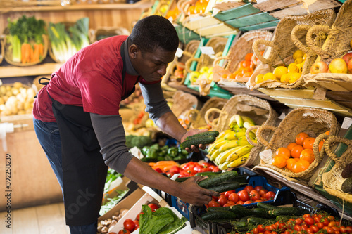 Confident African American seller in apron arranging fresh vegetables and fruits on greengrocery counter..