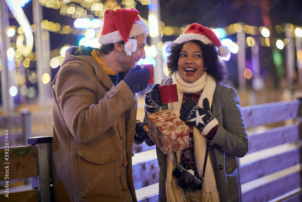 multiethnic couple talking outdoor on the street with santa hats, celebrating christmas, drinking tea