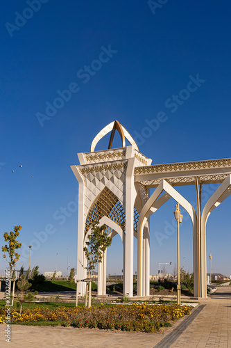 Zhibek Zholy park. The huge white gates of the park. Entrance. New park in the city of Turkestan. Park exterior design and decoration elements. Lawn and flowers. Blue sky. Lanterns as design elements 