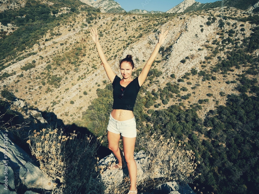 A smiling girl in shorts and a black T-shirt stands among the mountains with her hands up A smiling girl in shorts and a black T-shirt stands among the mountains with her hands up