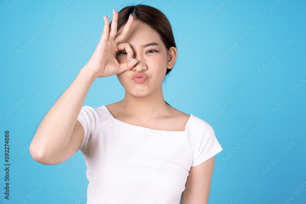 Asian woman looking at the camera through fingers in okay gesture, isolated on a blue background. Asian woman looking at the camera through fingers in okay gesture, isolated on a blue background.