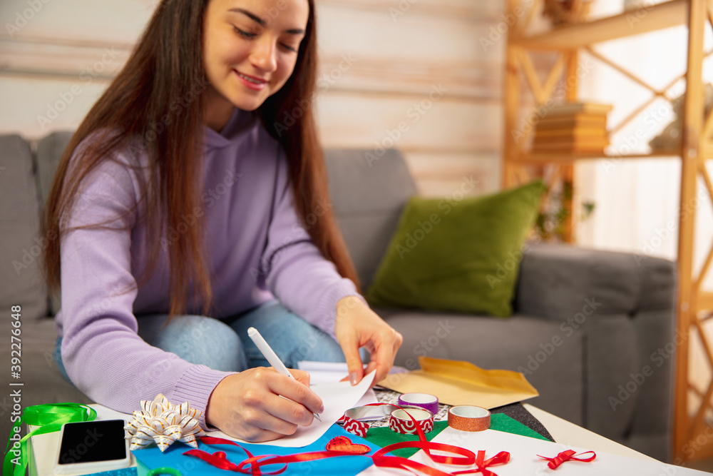 Fototapeta premium Comfortable. Woman writing letter, greeting card for New Year and Christmas 2021 for friends or family sitting at home with laptop. Holidays, celebration. Looks cheerful, happy, careful.