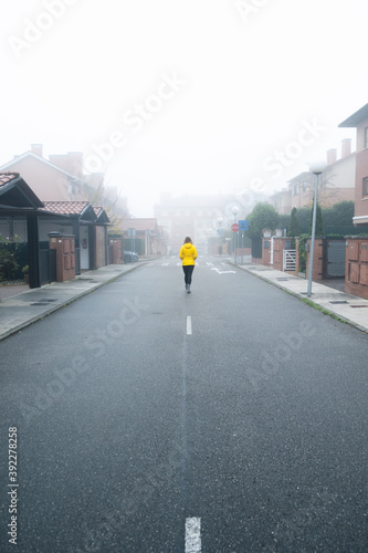 Young woman walking towards the fog in the middle of the street wearing a yellow down jacket.