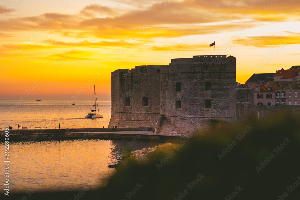 Golden sunset in the town of Dubrovnik, castle standing on the shore of a small harbour, sailboat leaving the harbour and a small local fishing boat entering. Tranquil simple serene setting