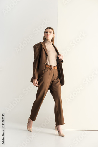 full length of young blonde woman in stylish brown suit walking on white photo