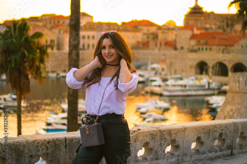 Fashionable attractive brunette posing on the city walls of Dubrovnik city as the sun sets during the golden hour. Small harbour in the distance illuminated by beautiful bright orange light