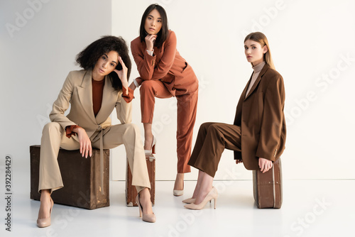 full length of stylish multicultural women in suits sitting on suitcases on white