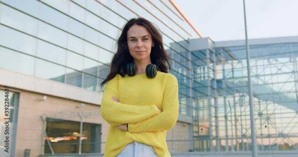 Front view of gorgeous smiling stylish young woman in trendy clothes which looking into camera with crossed arms near big urban modern building Front view of gorgeous smiling stylish young woman in trendy clothes which looking into camera with crossed arms near big urban modern building