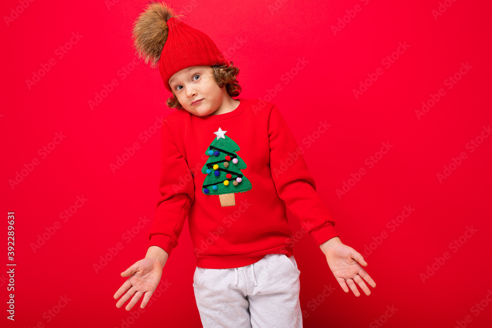 cool blond kid in warm hat and sweater with christmas tree on red background fooling around, christmas concept