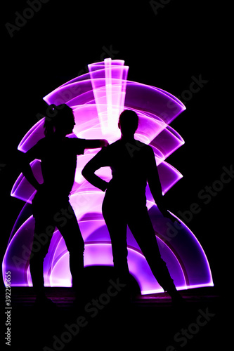 Lightpainting scene with girls. Silhouette of two women in front. Girl leaning on the shoulder of her friend. Abstract curved shape, pink color with a light saber in the background.