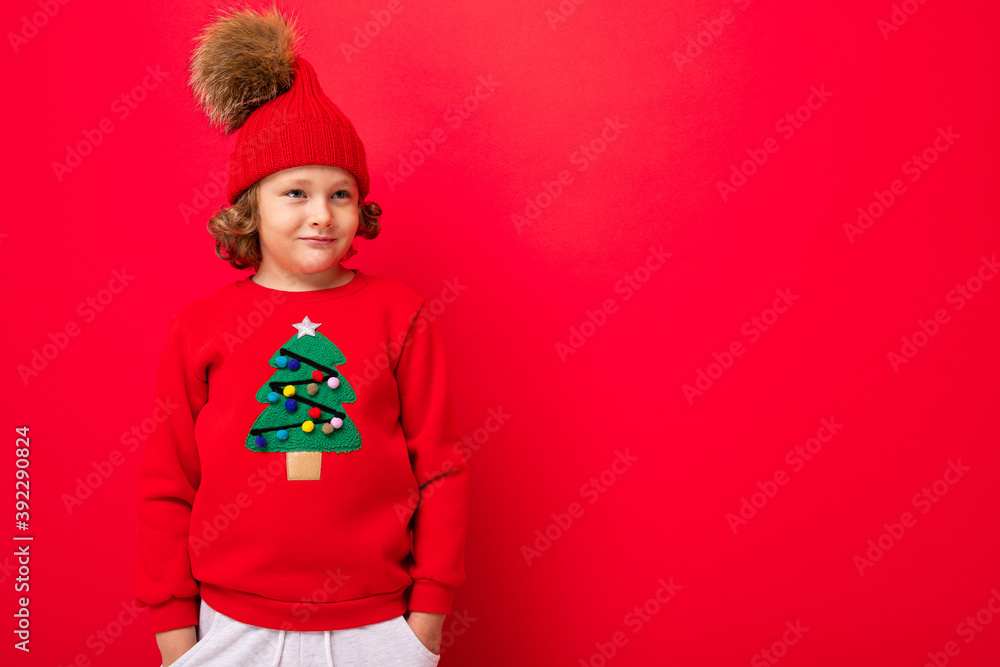 cute blond boy in warm hat and christmas sweater on red background with smile on his face