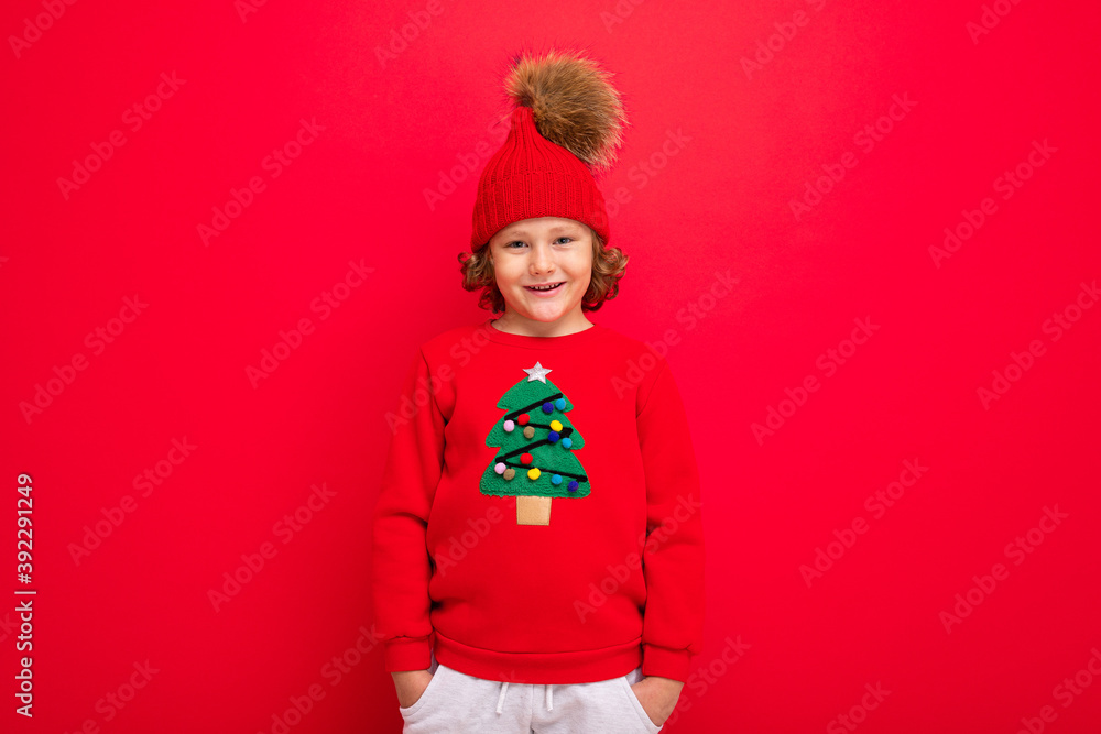 cute blond boy in warm hat and christmas sweater on red background with smile on his face